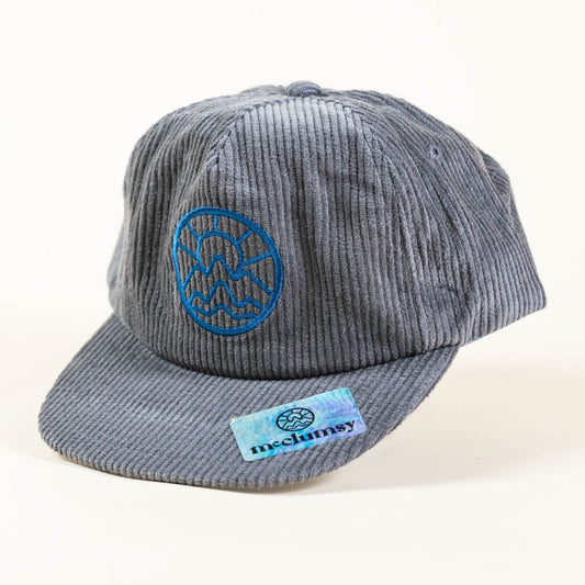 An unstructured, gray corduroy trucker hat with a flat visor, featuring a blue McClumsy embroidered logo on the front and a holographic rectangular McClumsy logo tag on the brim.